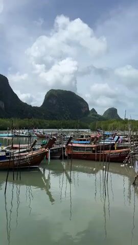 The video displays a serene waterfront scene with several longtail boats docked in calm, greenish water. In the background, lush green mountains rise majestically, their slopes covered in dense vegetation. The sky above is a mix of blue and white clouds, with some clouds casting shadows on the water. The boats, painted in shades of brown and blue, are tethered to wooden poles that jut out of the water. Some of the boats have people on them, seemingly preparing for or returning from a journey. The overall atmosphere is tranquil and picturesque, capturing the essence of a peaceful coastal landscape.
