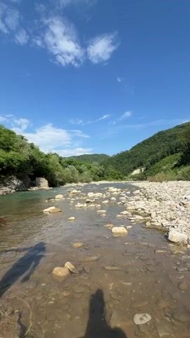 The video shows a shallow, clear river flowing through a valley. The riverbed is rocky, with many light-colored stones visible beneath the water. On the left bank, lush green trees and vegetation grow, with a stone structure partially visible. The right bank is also covered in green foliage, sloping upwards into forested hills. In the distance, more hills covered in trees stretch towards the horizon. The sky is a vibrant blue with a few white, wispy clouds. The perspective is low, close to the water's surface, and two shadowy figures, presumably of people, are cast on the riverbed in the foreground. The overall scene depicts a serene, natural environment on a sunny day.