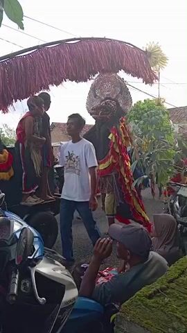 The video shows a traditional Indonesian dance or cultural performance taking place on a village street. Several people stand around watching, while others perform in colorful costumes. One notable figure is wearing an elaborate mask and costume in red, yellow, and black, suggesting a mythical or traditional character. There is also a decorative fringe overhead, adding to the festive atmosphere. Motorcycles parked along the side of the road indicate this is a public gathering or parade in a residential area. The sky is overcast.