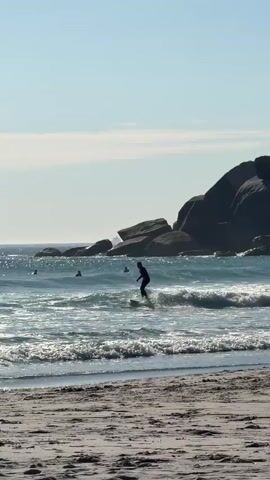 The video shows a sandy beach on a sunny day. In the foreground, the beach is mostly flat with scattered impressions in the sand. The ocean stretches out behind the beach, with gentle waves breaking near the shore. Several surfers, clad in dark wetsuits, are visible in the water. One surfer closer to the shore is actively riding a small wave. In the background, a cluster of large, dark rocks rises out of the water, partially obscuring the horizon. The sky is a pale blue with a few scattered clouds.