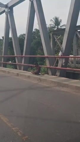 The video shows a person sitting on the side of a bridge, facing away from the camera. The person is wearing dark clothing and appears to be looking down. The bridge has a metal railing and a concrete barrier. In the background, there are trees and a cloudy sky. The overall scene is somewhat desolate.