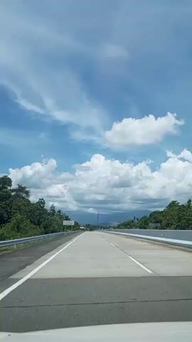 A vertical video shows a clear view of an empty, multi-lane highway stretching into the distance. The sky is blue with scattered white clouds. Lush green trees line both sides of the highway. A metal guardrail is visible on the left side of the road. A white sign with some text is positioned along the left side. The road surface appears to be made of concrete with white lane markings. The video appears to be filmed from a moving vehicle, looking straight ahead.