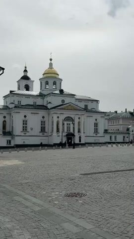 The video pans across a large cobblestone square in front of a white church with a gold dome and a smaller, mostly white building. Both buildings are surrounded by fences made of short, grey posts. In the background, there are more white and light-colored buildings. Some people walk around the square. The sky is overcast and grey.