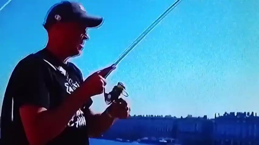The video shows two men fishing. The first man, wearing a black t-shirt and a blue baseball cap, is holding a fishing rod and reel. The second man, wearing a black fishing suit and a red life vest, is standing in shallow water, holding a fishing rod. The third clip shows a close-up of the second man's legs in the water. The fourth clip shows the two men talking to each other. The environment appears to be a lake or river with tall reeds.