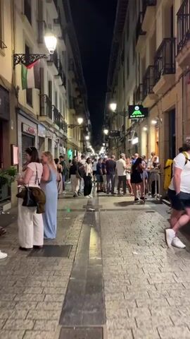 A narrow cobblestone street at night is lined with buildings on both sides. The buildings have multiple stories with balconies and windows. Streetlights illuminate the street, casting a warm glow. Several people are walking and standing in the street, some gathered in small groups. One group of people is standing outside a bar or restaurant, with tables and chairs set up. The people are dressed in casual clothing. The overall atmosphere appears to be lively and social. There are flags hanging from some of the buildings.