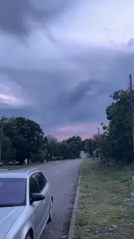 The video presents a static, eye-level shot of a rural or semi-rural road scene, likely in Kosten, Bulgaria. In the foreground, on the left, a silver Audi station wagon is parked on the side of an asphalt road. The road stretches into the distance, lined by lush green trees on both sides. The sky is covered with heavy, dark grey clouds, but a distinct band of pink and purple light is visible on the horizon, indicating either sunset or twilight. Utility poles stand along the roadside, particularly on the right. On the left, behind the parked car, a few benches are visible beneath the trees. The overall atmosphere is calm and serene. In the background, non-English vocal music, possibly a prayer or chant, can be heard. No explicit activity or visible text is present in the video.