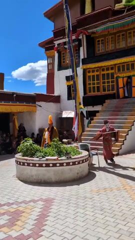 A group of people are gathered in an outdoor courtyard. In the center of the courtyard is a circular, raised planter with green plants. Two individuals are performing a ritual or dance. One individual, dressed in a red and gold robe and a white mask, moves towards the planter, holding a long, thin object. Another individual, wearing a yellow headdress and a yellow and gold robe, stands near the planter, holding a white object. The courtyard is paved with colorful tiles. In the background, there is a multi-story building with red and white walls and yellow-trimmed windows. Several people are seated on the ground to the left, watching the performance. A flag pole with several colorful flags stands behind the planter. The sky is blue with scattered clouds.
