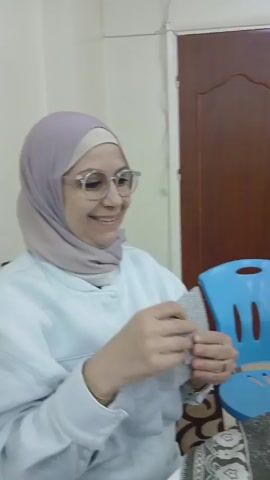 The video shows a woman in a black hijab playing cards at a table. She is smiling and seems to be enjoying the game. There are several cards laid out on the table. The room appears to be a living room or dining room. There is a door in the background. There is a chair to the left of the table. The tablecloth has a pattern on it. The woman is looking at the camera and laughing.