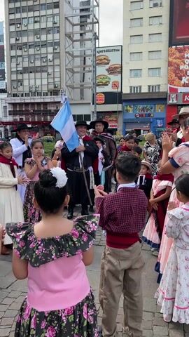 The video shows a group of people, primarily children, gathered in a public space. The children are dressed in traditional clothing, including floral dresses and checkered shirts with beige pants. They stand in a circle, clapping and cheering.  An older man stands in the center holding an Argentinian flag. Several adults are also present, some wearing traditional attire with hats and capes. They also participate in the clapping. The group is standing on a brick-paved area. Behind the group are tall buildings with various advertisements, including King’s Selection and KFC. The atmosphere appears celebratory and patriotic.