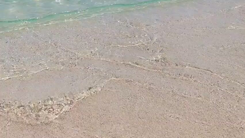 A close-up, high-angle shot shows gentle turquoise waves lapping onto a sandy beach. The water is clear, revealing the light-colored sand beneath, which has some darker patches and small indentations. The sunlight creates shimmering patterns on the water's surface. The camera angle remains static throughout the clip.