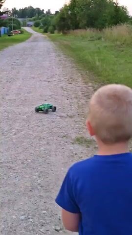 The video shows a young boy standing on a dirt road as two remote-controlled cars drive away from him. The road is lined with grass and trees, and houses are visible in the distance. The sky is a soft pink and orange, suggesting it is either sunrise or sunset. The boy is wearing a blue shirt and camouflage shorts, and yellow sandals. One of the cars is green and the other is black. The video captures the cars moving along the road.