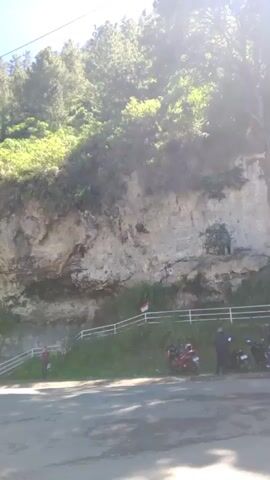 The video captures a scenic view of a steep, rocky cliff face covered in lush greenery, with trees densely populating the upper slopes. The bright sunlight suggests it is midday, and the clear blue sky indicates fair weather. A paved road runs along the base of the cliff, with a few motorcycles parked on the side, hinting at a tourist or recreational area.

A white railing lines a path that ascends the cliff face, with stairs visible in several sections. A small, reddish-brown, open-sided structure, possibly a pavilion or shelter, is nestled among the trees on the left side of the frame. A flagpole with a flag, likely the Indonesian flag given the location context, is positioned on the cliff face, adding a touch of national pride to the landscape.

A few people are visible near the road and on the ascending path, suggesting that people are visiting or exploring the area. The overall atmosphere is one of natural beauty and tranquility, with the sounds of nature and distant human activity creating a peaceful ambiance. The video appears to be a casual recording of a beautiful outdoor location.