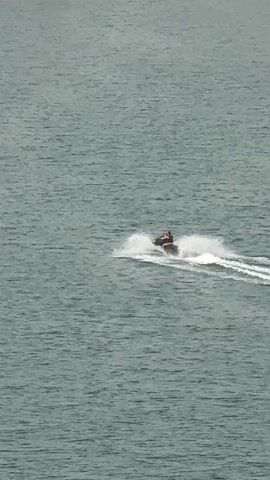 A jet ski is seen moving through the water from a high vantage point. The jet ski is dark in color and a single person in an orange life vest is riding it. The jet ski is creating a wake as it moves, leaving a trail of white spray behind it. The water is choppy and appears to be a grayish-blue color. In the distance, there is a blurry shoreline with some yellow and pink buoys. The video is shot from above and slightly to the side of the jet ski, providing a clear view of the action. The overall impression is of a sunny day with good conditions for water sports.