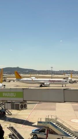 A Pegasus Airlines airplane, with "flypgs.com" written on its fuselage and the Pegasus logo on its tail, is parked at an airport gate under a clear blue sky. A jet bridge is connected to the airplane, with the word "PARIBU" visible on the bridge. In the foreground, a set of mobile stairs is positioned next to a white vehicle. The background shows other airplanes and airport infrastructure, including buildings and lights, with hills and distant buildings in the far distance. The scene appears to be a typical airport environment on a sunny day.