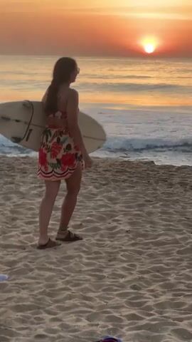 A young woman with dark hair pulled back in a ponytail walks across a sandy beach, carrying a surfboard under her right arm. She is wearing a floral bikini and a matching sarong. The sun is setting over the ocean, casting a warm, orange glow on the scene. The waves are gently rolling onto the shore. The woman turns to face the camera and smiles, then makes a peace sign with her fingers.