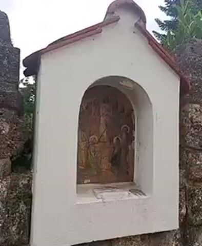 The video opens with a close-up shot of a small, white, shrine-like structure. The shrine has a red-tiled roof and an arched opening containing a religious painting. The painting depicts a scene with several figures, possibly a religious or historical depiction. The shrine is situated next to a large, rough-hewn stone structure, which appears to be part of a wall or an older building. The camera then pans to the right, revealing more of the stone structure and then moving further to show a glimpse of a house in the background, partially obscured by lush green bushes and trees. A decorative metal fence is visible in the foreground, separating the viewer from the property with the house. The sky appears overcast.