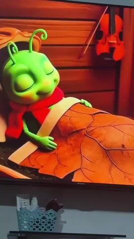 A 3D animated scene shows a small, green, cartoonish insect with large eyes and antennae sleeping in a bed made of a large, orange leaf. The insect is wearing a red scarf and is covered by a light-colored blanket. The bed is positioned within a hollowed-out nut shell, which sits on a surface covered with dry grass. In the background, a wooden wall is visible, with a small red violin hanging on it. The scene has warm, autumnal colors.