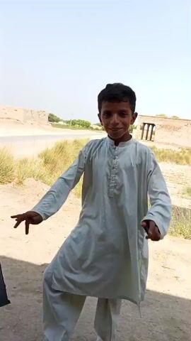 A video depicts two young boys outdoors, possibly in a rural area, under a bright sky. The boy in the foreground, wearing a light-colored shalwar kameez, strikes a martial arts pose with his hands raised. He is centered in the frame. In the background, another boy, dressed in a tan shalwar kameez, is walking and looking towards the camera. He is positioned to the right of the foreground boy. The ground is dusty and uneven, with some dry grass and what appears to be a brick visible on the ground. The environment suggests a dry, sunny climate with sparse vegetation and some simple structures in the distance.