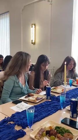 The scene shows women seated at a long table, with what looks to be a party, gathering, or dinner occurring. The women are of varying ages and appear to be conversing and enjoying a meal. They are using forks to eat. Each person has a square plate that seems to have an assortment of foods on it. The tables are adorned with a blue runner, glasses with a blue liquid in them, and lit candles. The room is brightly lit with natural light coming from a window on the left. There is also an ornate light fixture on the wall. The setting appears to be in a formal dining room or banquet hall. Cell phones are seen on the table in front of some of the women.