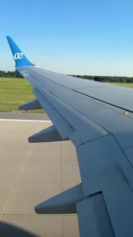 The video shows an airplane wing on a runway. The wing is gray and has a blue logo on the wingtip. The sky is blue and clear. The ground is green and grassy. The airplane is moving forward on the runway.