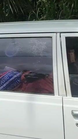The video shows a white Toyota Corolla station wagon driving on a road. The car has a license plate that reads "64-2842". The driver is a man with dark skin, wearing a blue shirt. The road is paved and has white lines painted on it. There are trees and buildings on either side of the road. The car is moving away from the camera.