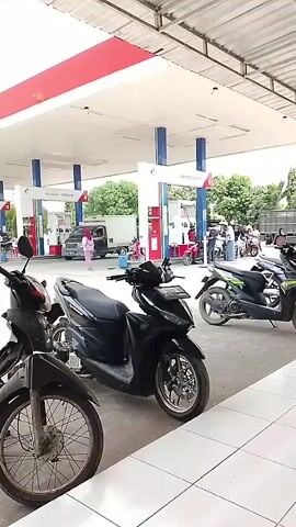 A still image shows a gas station with several motorcycles parked in the foreground. In the background, there are gas pumps, a truck, and a few people standing around. The gas station appears to be well-lit and operational. The overall scene is one of everyday activity at a public service location.