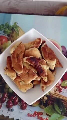 The video features a close-up overhead shot of a white, square-shaped bowl filled with golden-brown, irregularly shaped fried dough pastries, identified as Hvorost. The pastries exhibit varying shades of brown, suggesting different levels of crispness. The bowl is positioned on a vibrant tablecloth decorated with a detailed and colorful fruit pattern, including images of sliced watermelon, purple and green grapes, red cherries, kiwi slices, a melon, apples, and red currants. The background beyond the tablecloth is indistinct. An unseen speaker presents the dish, saying, "Hvorost for you. Look how beautiful it is." The scene suggests a homemade food presentation in a domestic setting, likely a kitchen or dining room.