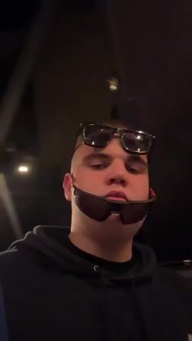 A young man is standing indoors wearing a black hooded sweatshirt and two pairs of sunglasses, one on his forehead and one around his chin. He moves the camera to the side, showing another young man in a black suit DJing. The camera returns to the first young man's face.