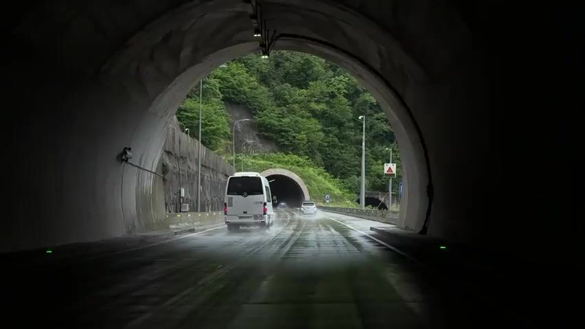The video begins inside a tunnel, looking out at a road that leads to another tunnel. It is raining. A white van and a silver car drive towards the second tunnel.  There is a rock wall on the left side of the road, and a guard rail and signage on the right. As the viewer exits the first tunnel, the camera pans up to reveal a lush green hillside on the right, covered in dense trees.  The peak of the mountain is shrouded in thick fog.