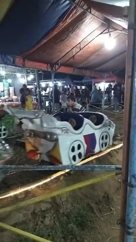 The video shows a small amusement park ride with colorful cars on a track. The cars are shaped like stylized vehicles and are moving along the track. Several people are visible, some riding in the cars and others standing around the ride. The area appears to be outdoors, possibly at a fair or carnival, with tents and lights in the background. The lighting suggests it is evening or nighttime. The ride seems to be in operation, with the cars moving smoothly. The overall atmosphere appears to be festive and casual.