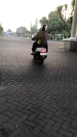 The media shows a person riding a scooter from behind. The person is wearing a helmet and a headscarf. The scooter is white and has a red taillight. The scene is an outdoor setting with a paved road. In the background, there are buildings, trees, and a mosque with a green dome. The time of day appears to be early morning or late afternoon, as indicated by the soft lighting and long shadows. The weather seems pleasant. The location cues suggest a place in Southeast Asia, possibly Indonesia, given the presence of a mosque and the typical architecture. No text is visible in the media.