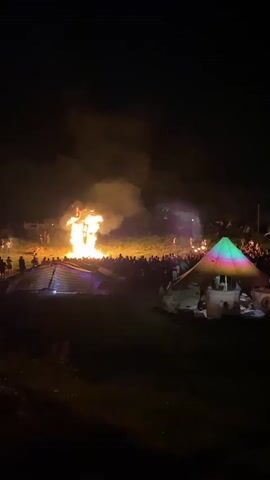 A large bonfire is burning in the center of the frame, illuminating the night sky with flames and smoke. A crowd of people are gathered around the fire, silhouetted against the light. Two tents or structures are visible in the foreground, one on the left with lights strung across it, and another on the right with colored lights projected onto its surface. The ground appears to be uneven and covered in grass. The overall scene depicts an outdoor gathering or festival at night.