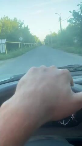 A first-person perspective video shows a person driving a car on a paved road. The driver's hand is visible resting on the steering wheel. The car's dashboard displays the time as 10:54, mileage as 21402, and the temperature as 8 degrees Celsius. The road is lined with trees and a fence on the left side. Overhead power lines are visible along the road. The sky is clear with some clouds. The video appears to be recorded during daylight.