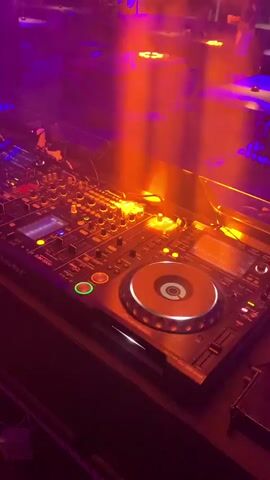 The video displays a DJ setup in a dimly lit club environment. The DJ equipment, a Pioneer DJM-2000 mixer and CDJ-2000s, is prominently featured. Lights, appearing as orange beams, illuminate the scene from above, creating a smoky atmosphere. In the background, blurred figures can be seen sitting at tables. The lighting shifts between orange and purple hues throughout the clip.