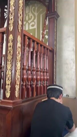 The video shows a man wearing a black shirt and a white and black prayer cap, facing away from the camera. He is seated or kneeling in front of an ornate wooden structure. The structure is dark brown, likely wood, and features intricate carvings and golden decorative elements on its pillars and railings. The railings are made of turned wooden spindles. Behind the wooden structure, a wall is visible with green and white decorative patterns. The lighting suggests an indoor setting, possibly a place of worship due to the man's attire and the context of the structure, which resembles a minbar or a prayer platform.