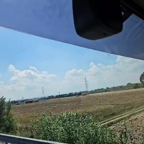 The video shows a view from inside a vehicle, looking out at a highway on a sunny day with a blue sky and scattered clouds. The highway curves to the left, with two lanes visible in the direction of travel. A metal guardrail runs alongside the highway. To the right of the highway, there is a field of dry vegetation, with some trees and utility poles in the distance. A small patch of greenery is visible on the left side of the road. A blue car is seen driving on the highway in the same direction as the camera. A dark object, possibly a dashboard camera, is mounted at the top of the windshield.