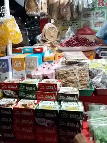 The video shows a street food stall, possibly in Indonesia, showcasing a variety of local snacks and sweets. The stall has multiple rows of packaged bakpia (a filled Chinese pastry), including the brand "Bakpia Pathok 024". There are also boxes of pie susu (milk pie) and pie tiramisu, plus cakes with pink frosting. In the background, we see dried goods hanging, a mound of red beans on a scale, and a vendor in a light blue shirt standing behind the counter. Two customers, likely children, are visible in the foreground, obscuring part of the stall's display.