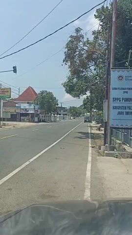 The video captures a street scene on a clear day, likely in Indonesia, based on the text on the sign. The camera is positioned inside a vehicle, looking forward. The road is paved and has clear lane markings. On the left side of the road, there are several small businesses with storefronts and awnings. A traffic light is visible in the distance. Further down the road, a motorcycle is being ridden. On the right side of the road, there is a sidewalk and a large blue sign with text and a government seal. Trees and utility poles line the right side of the street. The overall environment appears to be a suburban or semi-urban area with moderate traffic.