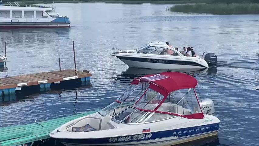A medium shot captures two boats docked and one boat moving on the water. In the foreground, a white Bayliner boat with a red canopy is docked. It has blue trim along its lower hull and a white outboard motor. A person can be seen inside the boat. To the left of this boat is a wooden dock with blue pontoons. In the middle ground, a white speed boat with a group of people on board is moving away from the dock. The boat has a license plate that reads "BB 0340 RUS 76". In the background, a large white and blue passenger boat is moving away from the shore. The water appears calm and the sky is overcast. The environment suggests a lake or river on a cloudy day, likely in a recreational area with a marina.