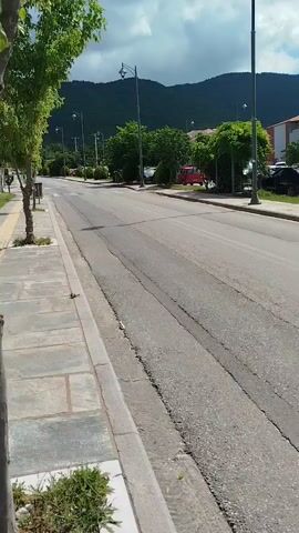 A video shows a street with buildings and cars. There are trees and mountains in the background. The street is paved and has a sidewalk. Several cars are parked along the street, and one car is driving away. A person is walking on the sidewalk. The sky is blue with some clouds.