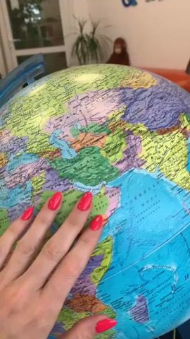 A close-up shot of a globe is shown. A person's hand with bright pink fingernails touches the globe. The globe displays continents and oceans with labels in Russian. In the background, a blurry figure wearing a brown outfit and a mask stands in front of a plant and a couch.