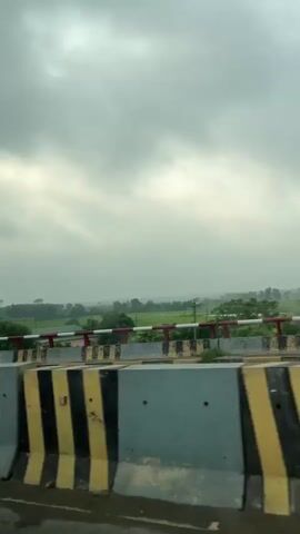 The video shows a view from a vehicle traveling on a road, likely on a bridge, as indicated by the concrete barriers with yellow and black diagonal stripes. Beyond the barriers, there is a railing with red and white horizontal stripes. The landscape is predominantly rural, with green fields and trees. The sky is overcast with gray clouds. A tall lamppost is visible on the left side of the frame, and a few distant buildings can be seen on the horizon. The road surface in the foreground appears wet, suggesting recent rain.