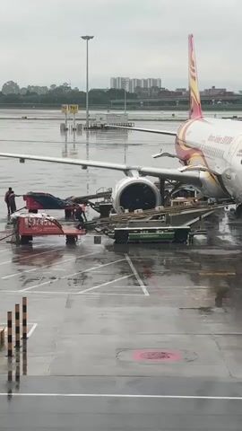 The video shows a rainy day at an airport. An airplane is parked on the tarmac, with ground crew working around it. Two individuals in orange vests are seen near a red baggage cart, one of whom appears to be handling a hose. Another individual in an orange vest is visible near a green luggage loader. The ground is wet and reflects the overcast sky and surrounding airport infrastructure, including lights and buildings in the distance.