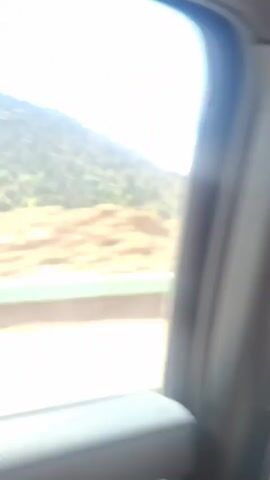 The video shows a view from a moving vehicle of a mountainous landscape. A green guardrail runs along the edge of the road. The mountains are covered in green trees and patches of brown rock. Further in the distance, the mountain peaks are covered in white snow and capped with clouds against a bright blue sky.