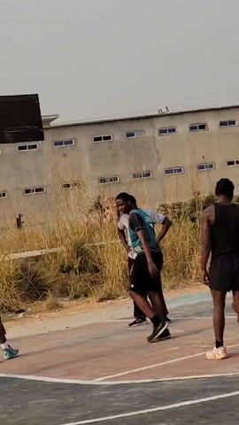 A video shows an outdoor basketball court with a group of people playing basketball. There are six males on the court. One is wearing a light gray t-shirt and dark pants, another has a green jersey and black shorts with the number 1 on the jersey, another wears a black tank top and shorts, and another has on a pink t-shirt with red shorts. Another male takes off his white shirt while standing on the court. One is dribbling the ball. Tall grass and a building with multiple small windows are behind the court. The sky is overcast.