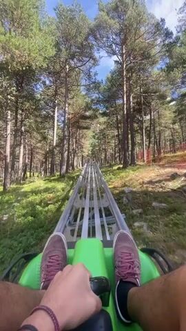 The video is a first-person perspective of someone riding a gravity-powered roller coaster through a forest. The rider's legs and hands are visible, gripping the handlebars of the green cart. The cart is on a metal track that winds through tall pine trees and lush green undergrowth. The sky is visible through the trees, with some clouds. The cart is moving at a moderate speed, and the camera is angled slightly downwards, capturing the track ahead and the surrounding scenery. There are no other people or carts visible in the immediate vicinity. The overall impression is one of a scenic and exhilarating ride through nature.