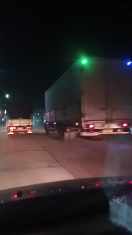 The video shows a nighttime view from a vehicle traveling on a road. In the foreground, the dashboard of the vehicle is visible. In the midground, two trucks are driving on the road. The truck on the right is larger and appears to be a semi-trailer truck. The truck on the left is smaller and has a covered bed. The road is paved, and there are streetlights and buildings visible on the sides of the road in the background. The sky is dark, indicating it is nighttime. The scene is somewhat blurry due to the movement of the vehicle and low light conditions.