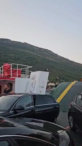 The video shows cars parked on a ferry with a large, green, ramp-like structure in the background. A wooded mountain is visible behind the ferry. The sky is clear and bright. One of the cars has its door open. A person can be seen through the window of the dark car on the right.