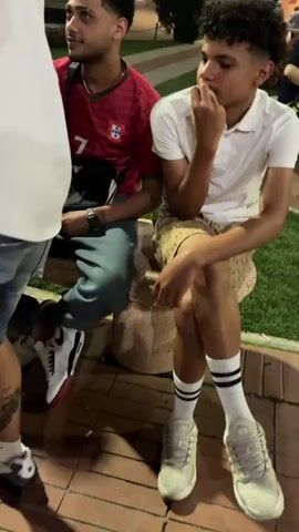 The video shows a group of people in a public space at night. Several individuals are wearing white T-shirts and shorts or jeans, and some are sitting down. A man in a red shirt is also visible. The camera moves through the crowd, focusing on different individuals. Buildings and streetlights are visible in the background.