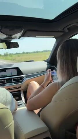 The video shows the interior of a car from the perspective of a passenger in the back seat. The camera is tilted upwards, showing a sunroof and the sky. In the front passenger seat, a young woman with blonde hair is looking at her phone. The driver is partially visible on the left, wearing a white hat. In the back seat, a person's legs are in the foreground, wearing white shorts with lace trim and a phone on their lap. Another person's legs are visible on the far right. The car appears to be on a road with fields on either side, and the sun is shining. The overall atmosphere suggests a road trip.